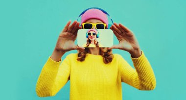 Kadının ellerini uzatıp kulaklıkla telefonuyla selfie çekerken mavi arka planda müzik dinlediği yakın plan.