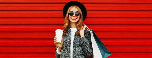 ショッピングバッグやグレーのコートを着てコーヒーカップと美しい笑顔の若い女性は カラフルな赤の背景に帽子 — ストック写真