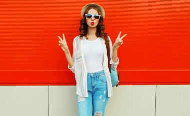 Portrait of beautiful young woman blowing her lips sending sweet air kiss posing wearing a summer straw hat on red background