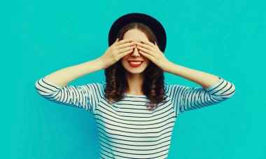 Portrait of happy surprised smiling young woman covering her eyes wearing a striped t-shirt, black round hat on blue background