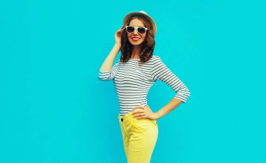 Portrait of happy smiling stylish young woman posing wearing a striped t-shirt and summer straw round hat on blue background