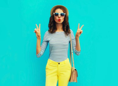 Portrait of beautiful young woman blowing her lips sending sweet air kiss posing wearing a handbag, striped t-shirt and summer straw round hat on blue background