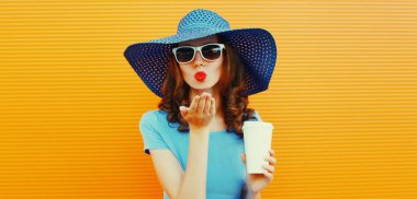 Portrait of beautiful young woman blowing her lips sending sweet air kiss posing wearing a summer straw hat, blue dress on orange background