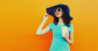 Portrait of beautiful happy smiling young woman with cup of coffee wearing a summer hat and dress looking away on city street over orange background