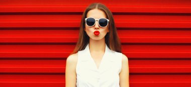 Kırmızı arka planda güneş gözlüğü takarak dudaklarını kırmızı rujla silen şık genç kadın modelinin portresi.