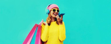 Şık, gülümseyen genç bir kadının renkli portresi. Alışveriş çantalarıyla arıyor kulaklıkla müzik dinliyor sarı örgü bir kazak giyiyor, mavi arka planda pembe şapka takıyor.