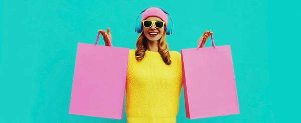 穿着黄色针织毛衣 蓝色背景的粉红帽子 头戴购物袋 戴着耳机听音乐的年轻女子多姿多彩的画像 — 图库照片