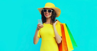 Akıllı telefonları ve alışveriş çantaları olan, renkli mavi arka planda sarı hasır şapka takan mutlu, genç bir kadının moda portresi.