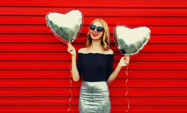 Kırmızı arka planda gümüş kalp şeklinde balonları olan güzel, mutlu, genç bir kadının portresi.