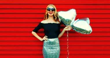 Kırmızı arka planda gümüş kalp şeklinde balonları olan güzel, mutlu, genç bir kadının portresi.