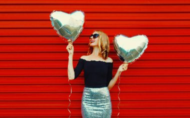 Kırmızı arka planda gümüş kalp şeklinde balonları olan güzel, mutlu, genç bir kadının portresi.