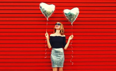 Kırmızı arka planda gümüş kalp şeklinde balonları olan güzel, mutlu, genç bir kadının portresi.