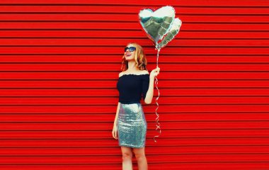 Kırmızı arka planda gümüş kalp şeklinde balonları olan güzel, mutlu, genç bir kadının portresi.