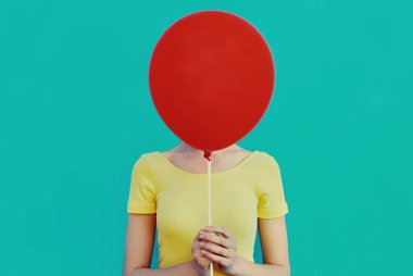 Kafasını mavi arka planda kırmızı balonla kapatan kadının portresi.