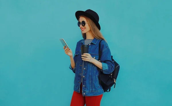 黒丸の帽子をかぶったスマートフォンを持つ笑顔の若い女性の肖像 青の背景にデニムジャケット — ストック写真