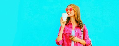 Akıllı telefonlu, pembe ceketli, mavi arka planda güneş gözlüklü, kahve içen şık genç bir kadının portresi.