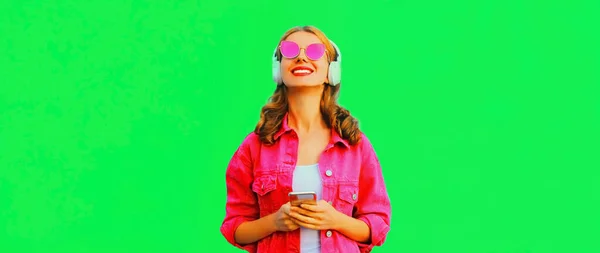 ピンクのジャケットを着て音楽を聞いてスマートフォンでヘッドフォンでスタイリッシュな幸せな笑顔の若い女性の肖像画 緑の背景にサングラス — ストック写真
