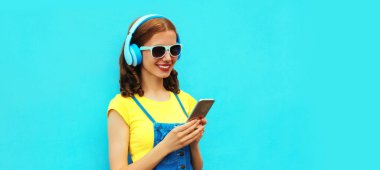 Mavi arka planda telefonla müzik dinleyen kulaklıklı mutlu genç bir kadının portresi.