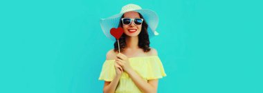 Mutlu gülümseyen genç bir kadının portresi. Kırmızı tatlı kalpli, lolipop şeklinde. Mavi arka planda yaz hasır şapkası takıyor.
