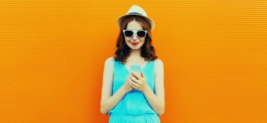 Turuncu arka planda yaz hasır şapkası takan akıllı telefonlu beyaz kadın portresi.