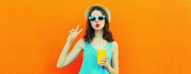 Taze meyve suyu içen genç bir kadının portresi. Turuncu arka planda yaz şapkası takıyor.