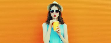 Taze meyve suyu içen genç bir kadının portresi. Turuncu arka planda yaz şapkası takıyor.