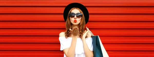 漂亮的年轻女子 戴着购物袋 嘴边飞扬着甜蜜的空气 亲吻着 戴着红色背景的黑色圆帽 — 图库照片