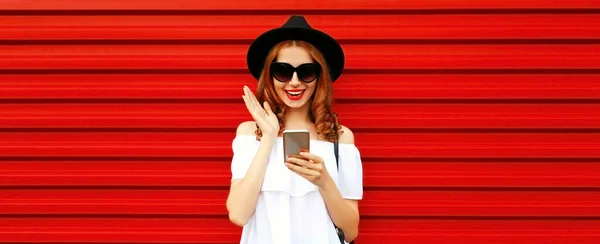 赤い背景にスマートフォンを持つスタイリッシュな幸せな笑顔の若い女性の肖像画 — ストック写真