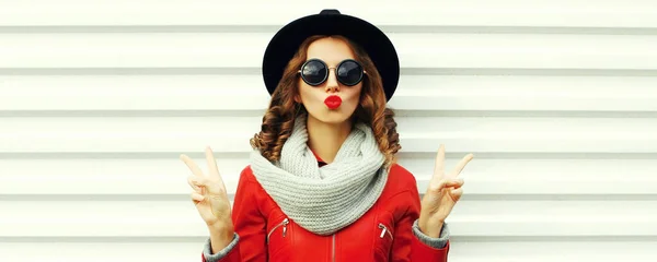 スタイリッシュな女性モデル吹いて彼女の唇送信甘い空気キス身に着けているスカーフと赤のジャケット 黒丸帽子上の白い背景 — ストック写真