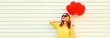 Sarı gömlek giyen, beyaz arka planda güneş gözlüğü takan, kırmızı kalp şeklinde balonları olan mutlu genç bir kadının portresi.