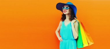 Renkli alışveriş çantaları olan, turuncu arka planda yaz hasır şapka takan, reklam metinleri için boş kopya alan güzel, mutlu genç bir kadının portresi.