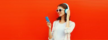 Kulaklıklı gülümseyen genç bir kadının portresi. Kırmızı arka planda telefonla müzik dinliyor.