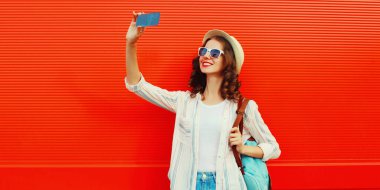 Mutlu gülümseyen genç bir kadının portresi telefonla selfie çekiyor yaz hasır şapkası takıyor, kırmızı arka planda sırt çantası takıyor.