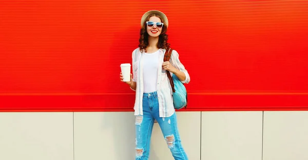 若い笑顔の女性は赤い背景の上に街の通りにバックパック夏のわら帽子を身に着けているコーヒーのカップで歩く 広告テキストのための空白のコピースペース — ストック写真