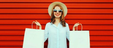 Renkli kırmızı arka planda hasır şapka ve gömlek giyen, alışveriş torbalarıyla gülümseyen genç bir kadının portresi.