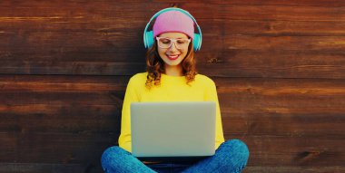 Modern mutlu gülümseyen genç bir kadının portresi dizüstü bilgisayarda çalışıyor ve şehir parkında kulaklıkla müzik dinliyor.