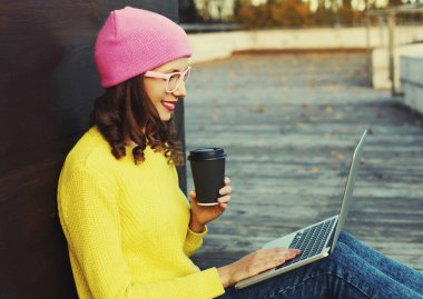 Şık, mutlu ve modern bir kadının portresi. Laptopuyla kahve içiyor. Renkli giysiler içinde şehir parkında oturuyor.