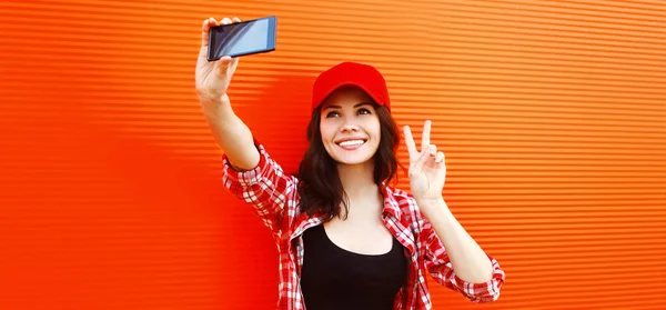 幸せな笑顔若い女性の夏の肖像写真によって自画撮りスマートフォン着用野球帽上の赤の背景 — ストック写真
