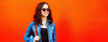 Mutlu gülümseyen genç bir kadının portresi. Renkli kırmızı arka planda sırt çantalı kot ceket giyiyor. Reklam metinleri için boş bir kopya alanı var.