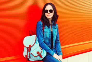 Genç esmer kadının portresi. Renkli kırmızı arka planda sırt çantalı kot ceket giyiyor.