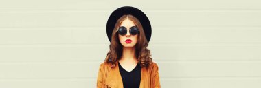 Siyah yuvarlak şapkalı, gri arka planda kahverengi ceketli, zarif genç bir kadının portresi.