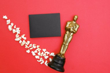 KHARKIV,, UKRAINE - JULY 1, 2020: Plastic Oscar award on a red background with popcorn and a black envelope. clipart