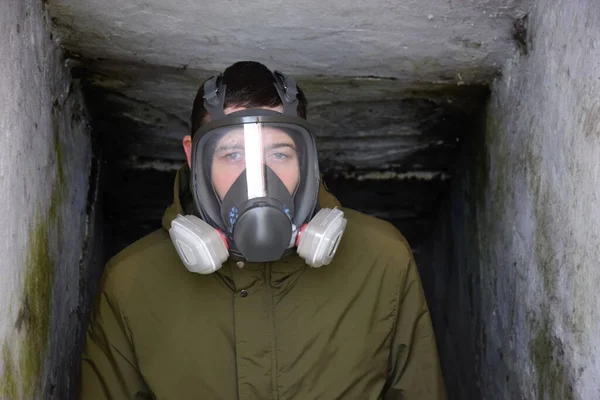 A young guy in a respirator in a bomb shelter. Dark photo. Personal safety from covid 19, toxic work, chemical weapons, nuclear threat.