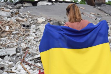 Bombalanmış bir okulun arka planına karşı Ukrayna 'nın ulusal bayrağı altında duran genç bir kız. Mavi ve sarı renkler. Ukrayna 'da Savaş.