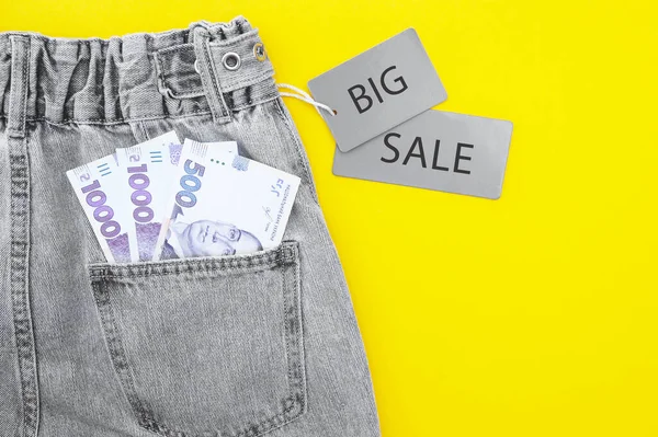 Flat lay, detail of denim jeans with Ukrainian hryvnia bills in pocket with gray label and inscription SALE on yellow background. Shopping and discount concept. Black Friday.