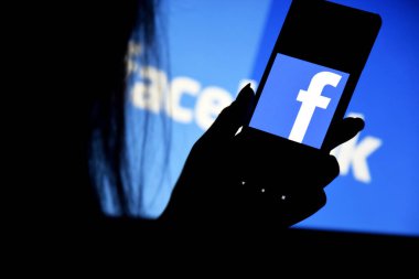 Kharkiv, Ukraine - Desember 20, 2021: A phone with a Facebook screen in the hands of a young girl against the background of a monitor. Selective focus.