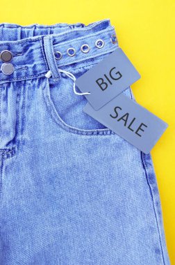 Flat lay, detail of denim jeans with gray label and inscription SALE on yellow background. Shopping and discount concept. Black Friday.