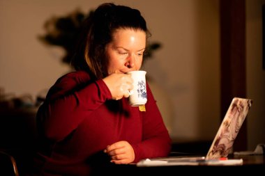 Evde bir fincan kahve ve dizüstü bilgisayarı olan genç bir kadın var.