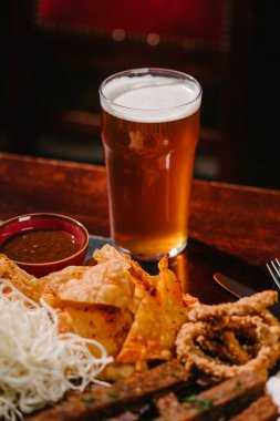 Tahtasında atıştırmalıklar olan bir bardak light bira. Patates ve pirinç cipsi, kızarmış ekmek, soğan halkaları, füme peynir, domates sosu ve tartar sosu.