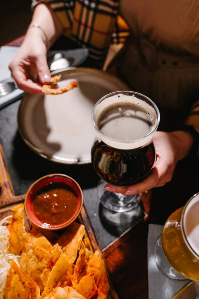 A girl drinks dark beer in a pub with beer snacks on a wooden board. Potato and rice chips, croutons, onion rings, smoked cheese, tomato sauce, tartar sauce.